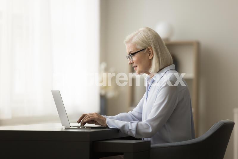 Positive busy senior business woman working from home, sitting at workplace table, typing on laptop computer at home, using pc for online job communication, wireless Internet connection