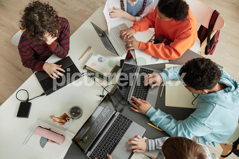 Top view at diverse group of teen school children using computers in classroom studying together at table