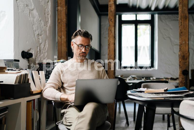 White middle-aged man in earphones using laptop computer while sitting in modern office