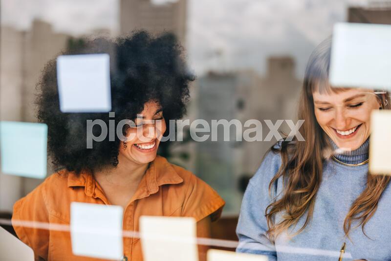 Cheerful young businesswomen brainstorming using sticky notes. Happy businesswomen discussing their business ideas during a meeting. Creative female entrepreneurs working as a team in a modern office.

