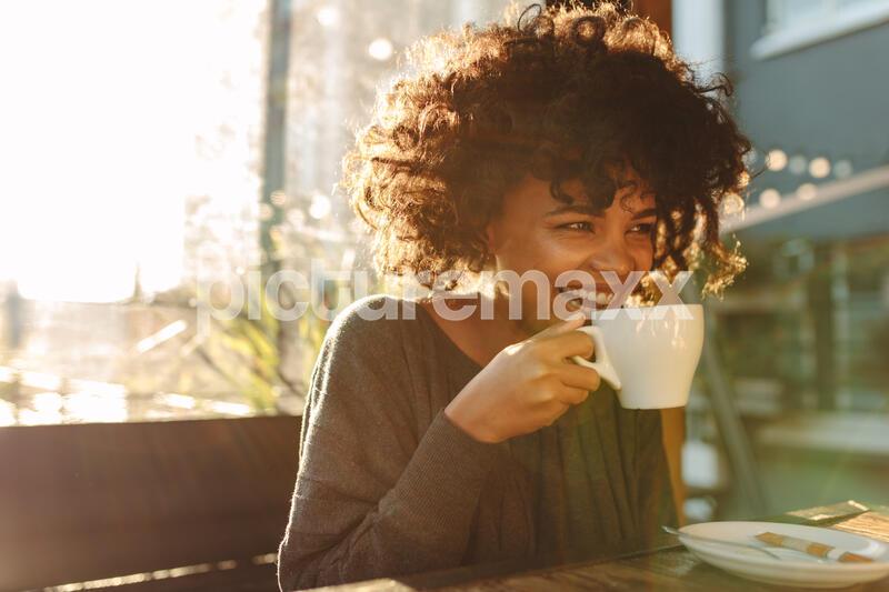 Smiling woman holding a coffee cup close to her face sitting inside a coffee shop. Curly haired woman drinking coffee.