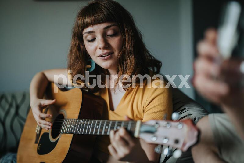 A young woman playing an acoustic guitar enjoys a peaceful moment in a cozy living room. The natural lighting adds warmth and relaxation to the scene.